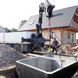 Hydrotech Tomasz Zglinicki - Montaż betonowego zbiornika za pomocą dźwigu samochodowego; w tle drewniany dom z balkonem i sterta żwiru.