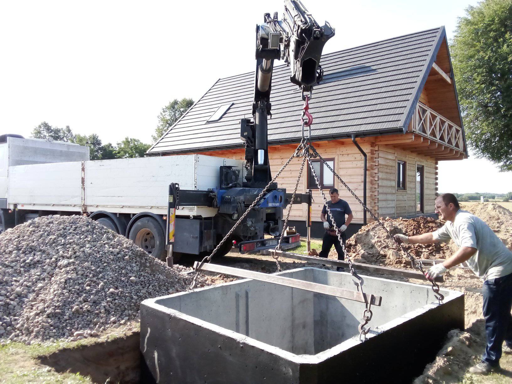 Montaż betonowego zbiornika za pomocą dźwigu samochodowego; w tle drewniany dom z balkonem i sterta żwiru.
