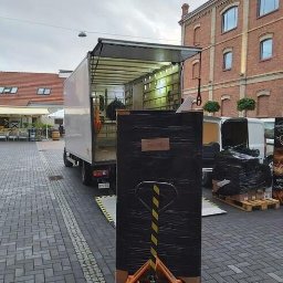 ANATOLII KUCHEREVSKYI - Duży karton owinięty czarną folią na wózku paletowym przed otwartą paką białej ciężarówki, rampa wyładowcza, kostka brukowa, budynek z czerwonej cegły w tle.