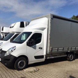 ANATOLII KUCHEREVSKYI - Biały samochód dostawczy Renault Master z plandeką na szarym brukowanym podłożu, zaparkowany obok innych pojazdów dostawczych pod błękitnym niebem.