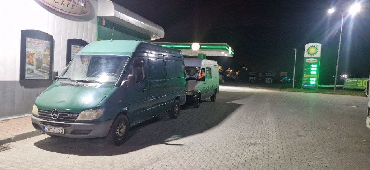 Dwa busy Mercedes-Benz Sprinter w odcieniach zieleni zaparkowane na stacji benzynowej BP nocą, z widocznym cennikiem paliw i szyldem kawiarni w tle.