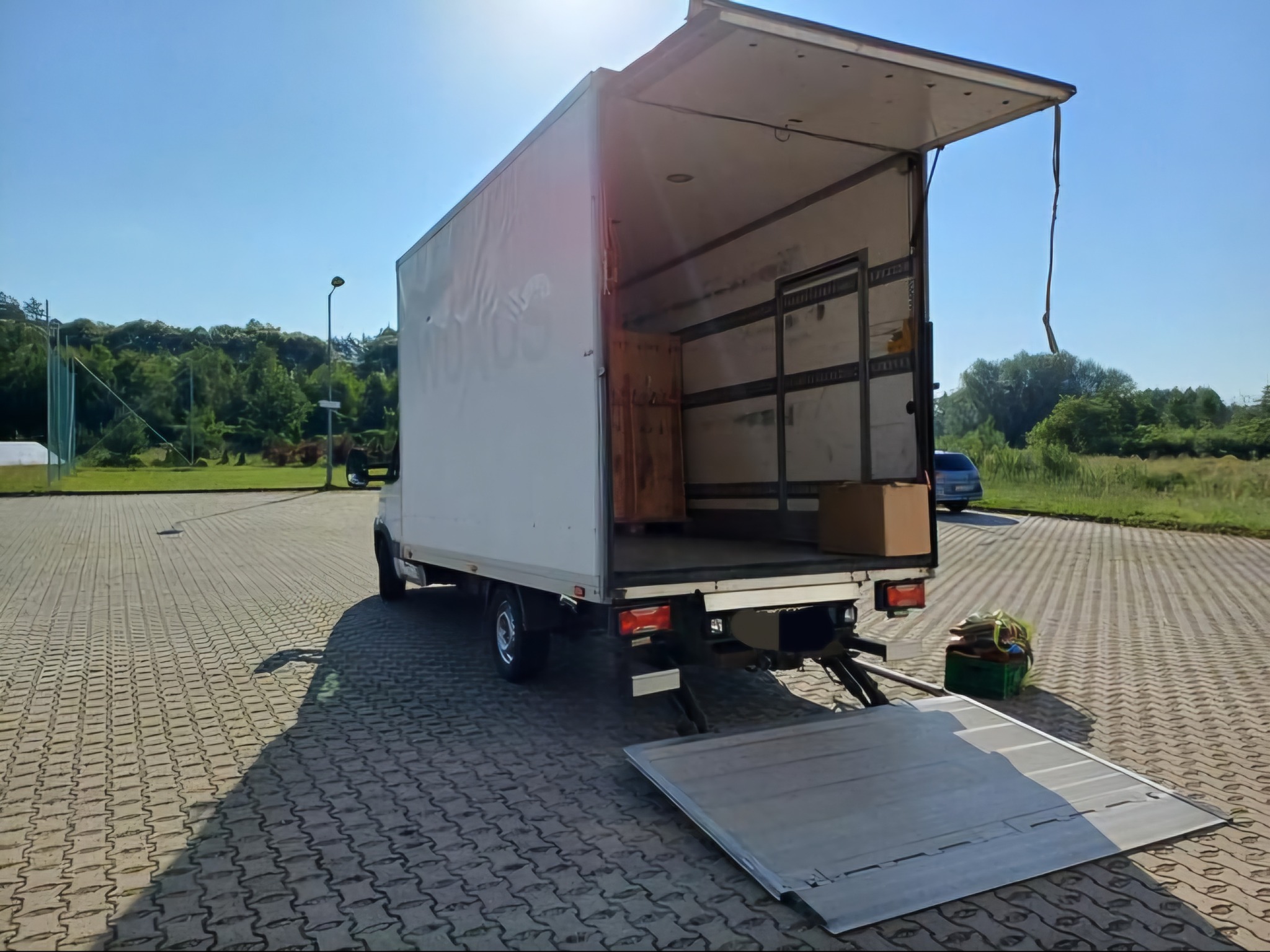 Otwarty tył białej ciężarówki z rozłożoną rampą na brukowanym parkingu w słoneczny dzień, widoczne kartony wewnątrz i zielony pojemnik obok rampy.