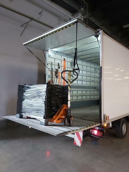 Paleta owinięta folią stretch, umieszczona na podnośniku paletowym, na platformie załadunkowej otwartego busa dostawczego z widocznymi pasami mocującymi wewnątrz.