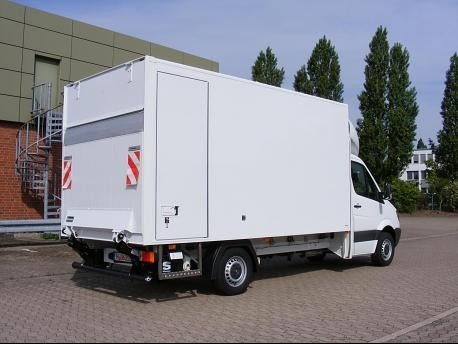 Biały samochód dostawczy typu furgon z zabudową kontenerową, widoczny z tyłu, zaparkowany na betonowym placu przed budynkiem z zieloną elewacją i spiralnymi schodami przeciwpożarowymi.