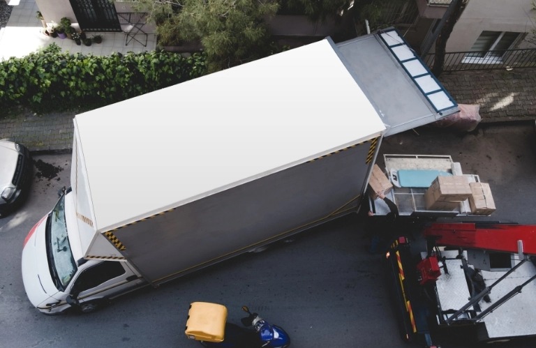 Widok z góry na białą ciężarówkę z otwartą przestrzenią ładunkową, z której wyładowywane są kartony i meble na platformę podnośnika, zaparkowaną na ulicy w otoczeniu budynków mieszkalnych i zieleni.