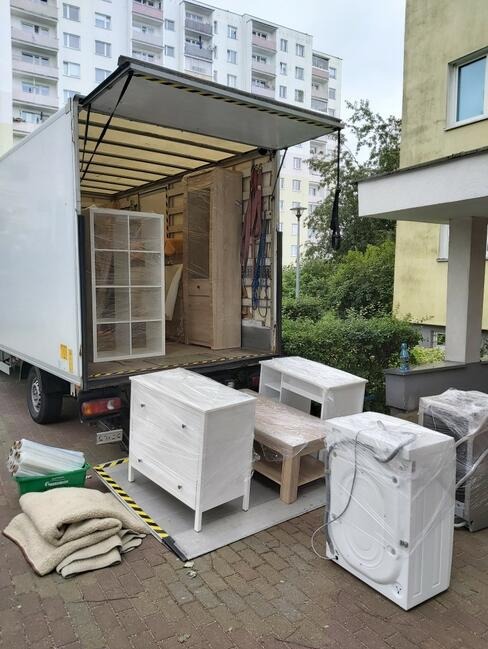 Załadunek mebli, zapakowanych w folię ochronną, do samochodu dostawczego w Bielsku-Białej. Widoczne komoda, regał, stolik i pralka, zabezpieczone koce i folia.