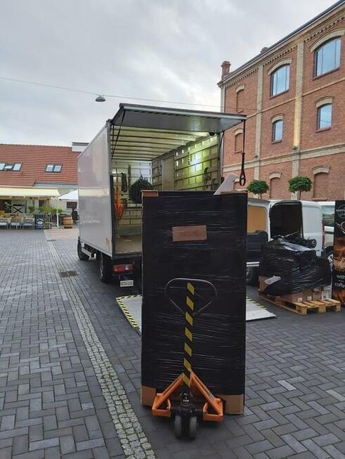 Duży karton owinięty czarną folią na wózku paletowym przed otwartą paką białej ciężarówki, rampa wyładowcza, kostka brukowa, budynek z czerwonej cegły w tle.