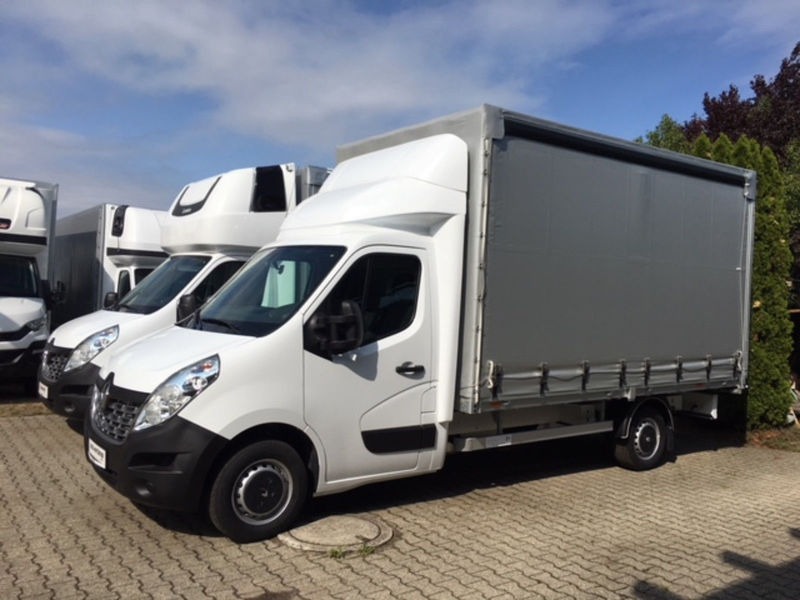 Biały samochód dostawczy Renault Master z plandeką na szarym brukowanym podłożu, zaparkowany obok innych pojazdów dostawczych pod błękitnym niebem.