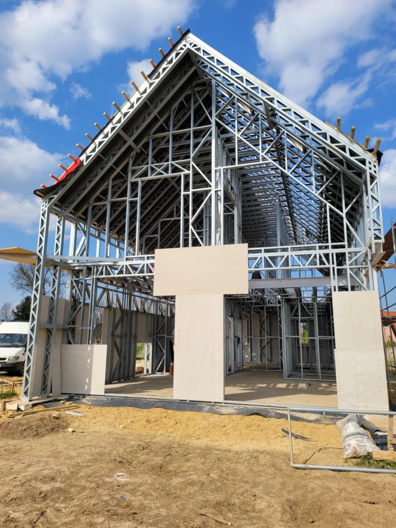 Konstrukcja szkieletowa domu jednorodzinnego w trakcie budowy, widoczne stalowe profile i częściowo zamontowane płyty elewacyjne, na pierwszym planie teren budowy.