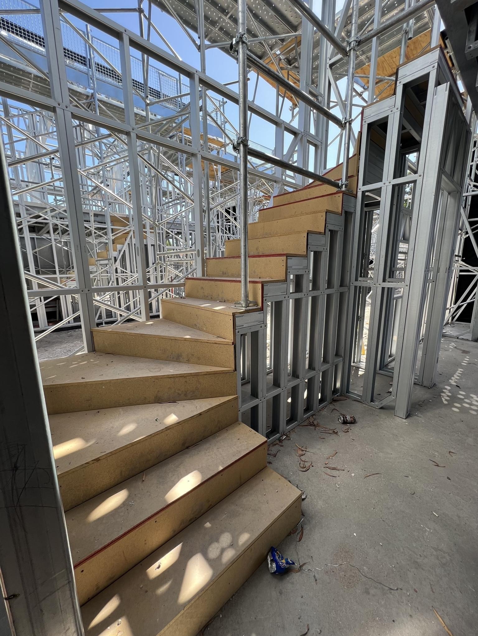 Konstrukcja domu szkieletowego w trakcie budowy, widoczne stalowe ramy ścian i drewniane schody tymczasowe, z bliska na surowym betonie.