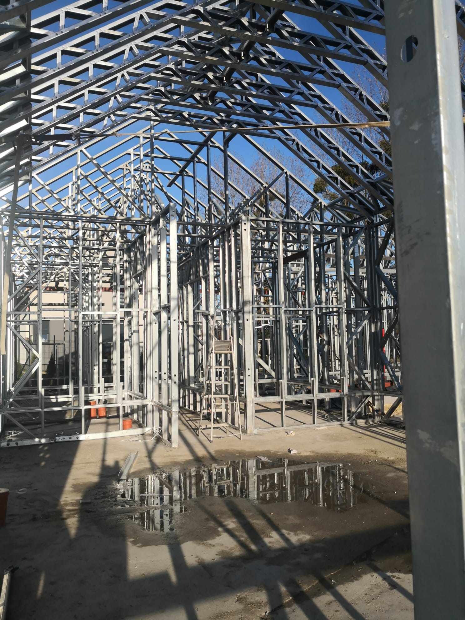 Metalowy szkielet domu w trakcie budowy, widoczne elementy konstrukcyjne, drabina i kałuża z odbiciem, błękitne niebo w tle.