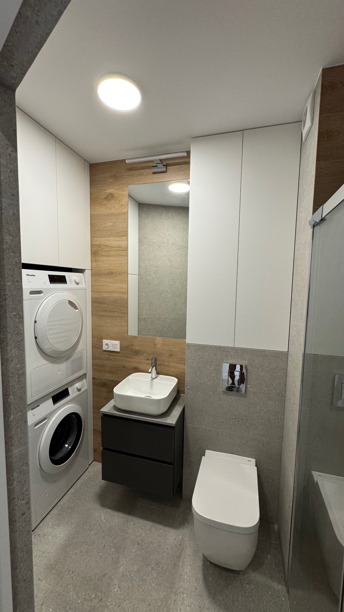 Nowoczesna łazienka z pralką i suszarką Miele w zabudowie, umywalką nablatową na czarnej szafce, podwieszaną miską WC i drewnopodobną ścianą za lustrem.