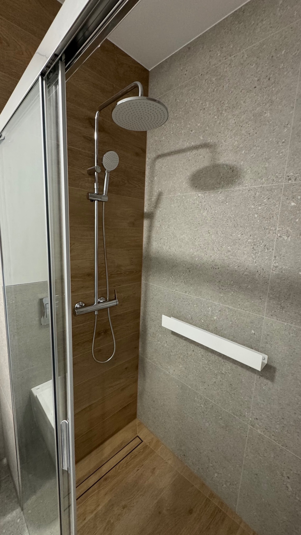 Nowoczesna kabina prysznicowa walk-in z deszczownicą i odpływem liniowym w podłodze, wykończona płytkami imitującymi drewno i szarym kamieniem.