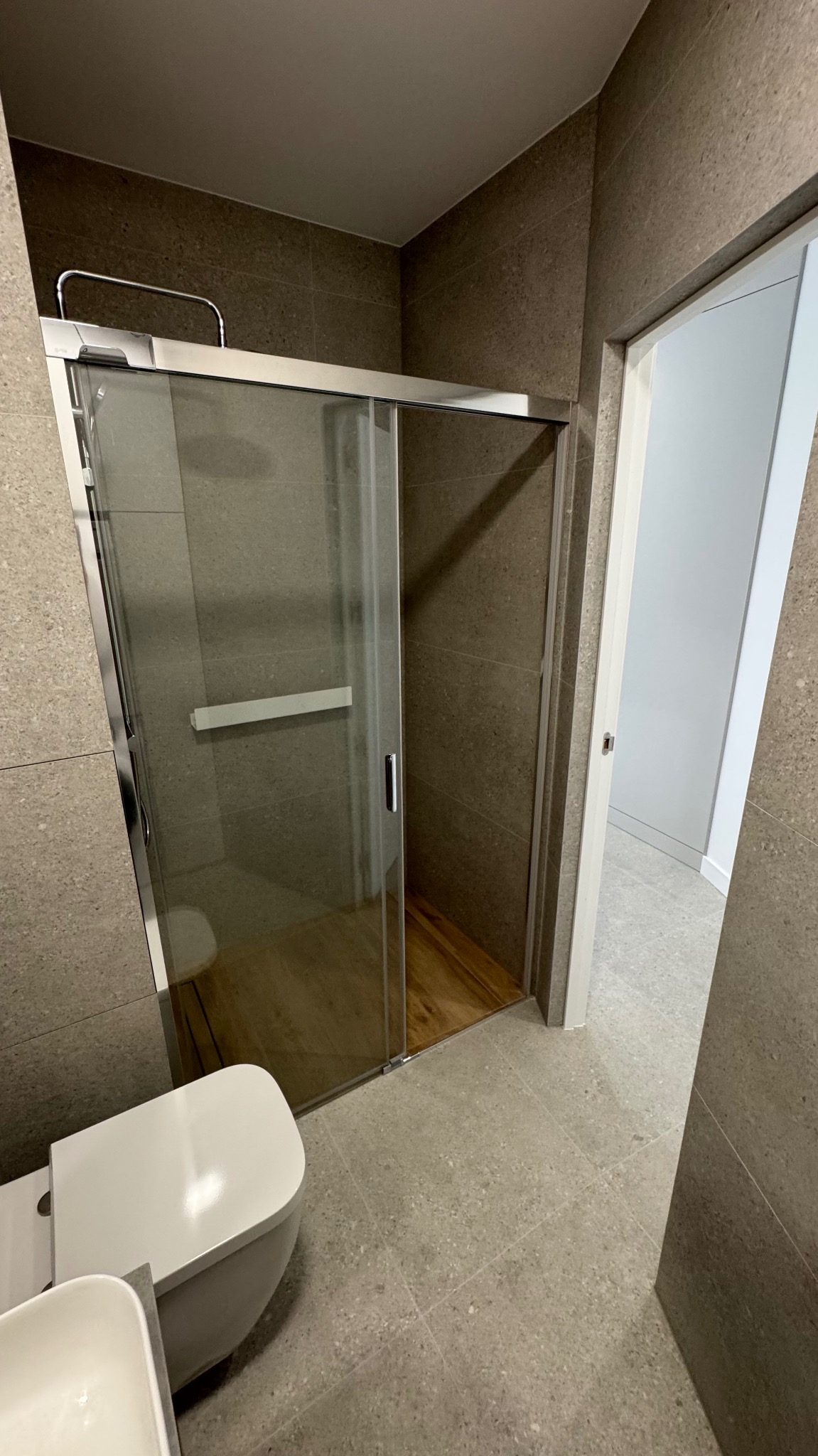 Nowoczesna łazienka z kabiną prysznicową z przesuwnymi drzwiami, wykończona płytkami w odcieniach szarości i brązu, z drewnianą podłogą w kabinie i częściowo widoczną toaletą.