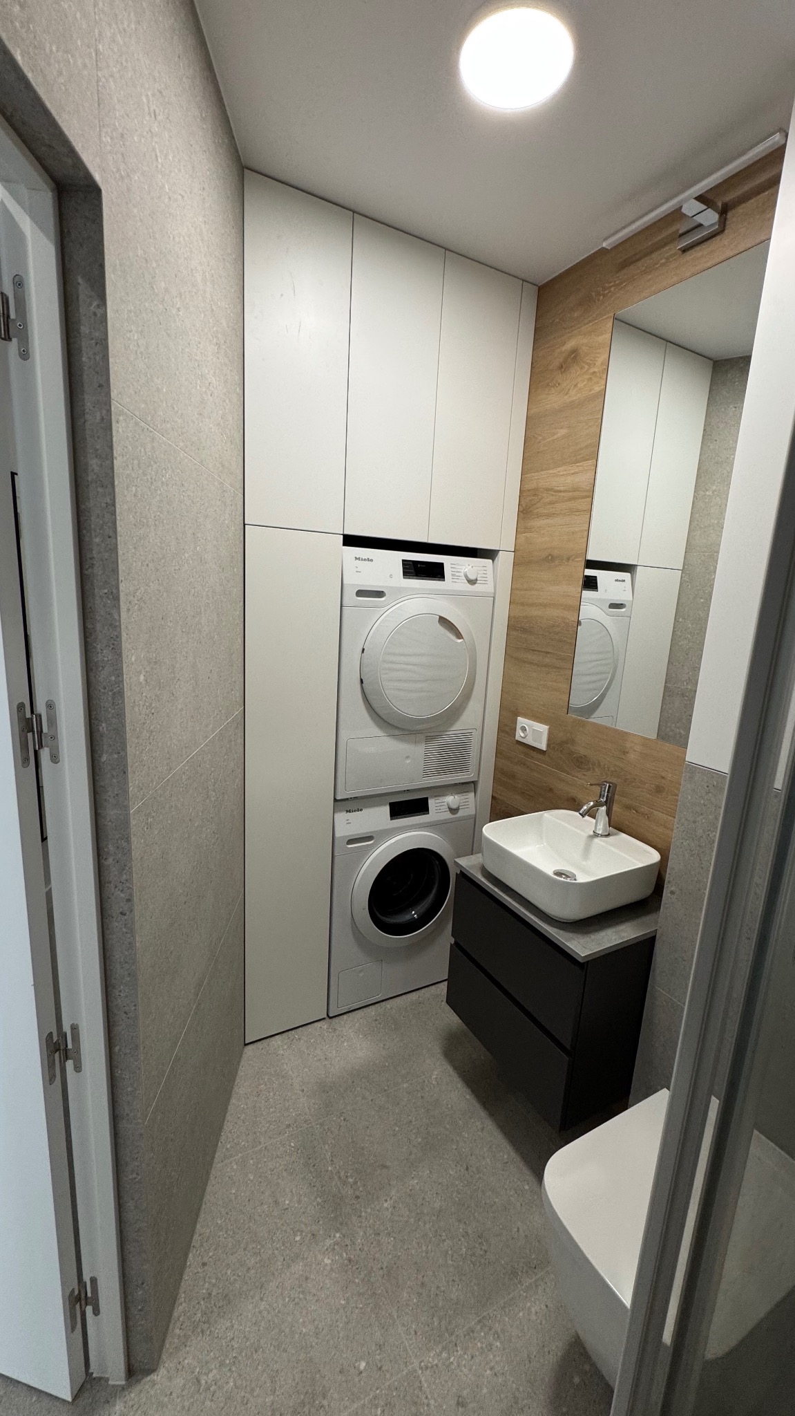 Nowoczesna łazienka w szarościach z pralko-suszarką Miele w zabudowie, umywalką nablatową na czarnej szafce i lustrem z drewnopodobną okładziną.