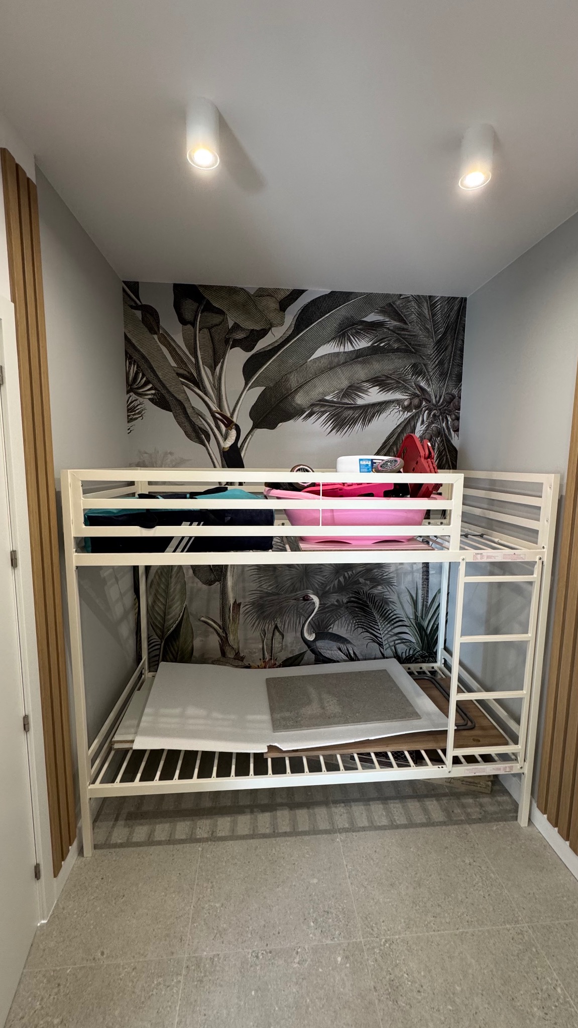 Białe łóżko piętrowe w pokoju z szaro-białą tapetą w motywy tropikalne, widoczne przygotowane płyty do wykończenia, podłoga wyłożona szarymi płytkami.