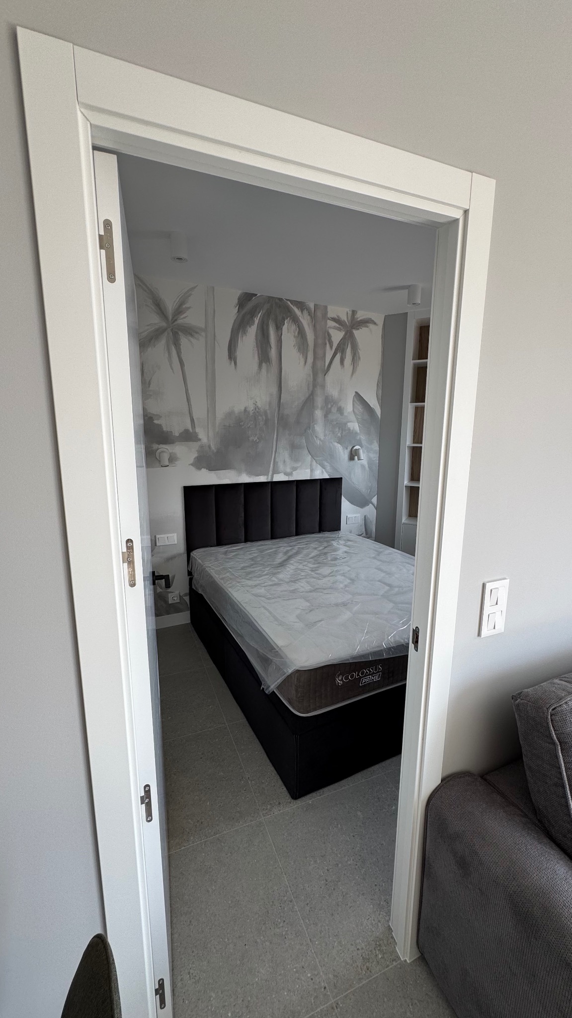 Widok przez białe drzwi na sypialnię z czarnym łóżkiem Colossus Prime z zagłówkiem i tapetą w palmy, łóżko zabezpieczone folią ochronną.