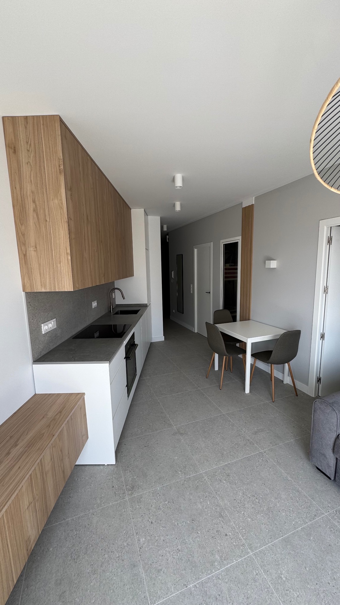 Nowoczesna kuchnia z drewnianymi szafkami górnymi, szarym blatem i płytkami podłogowymi w odcieniach szarości, widok na stół jadalny z krzesłami.