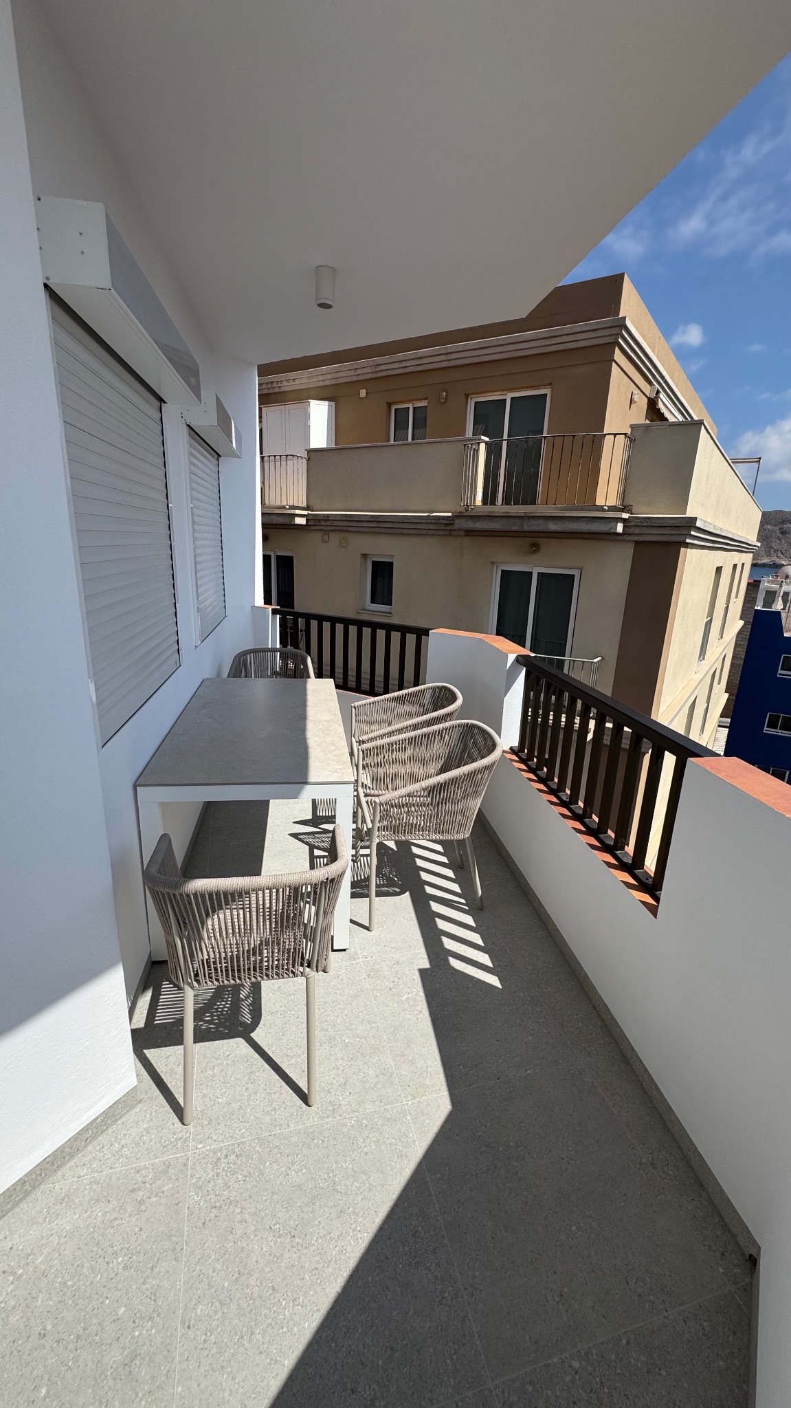 Jasny balkon z szarymi płytkami, białymi ścianami i zestawem mebli ogrodowych: stół i cztery plecione krzesła. W tle widoczny budynek z balkonami i niebieskie niebo.