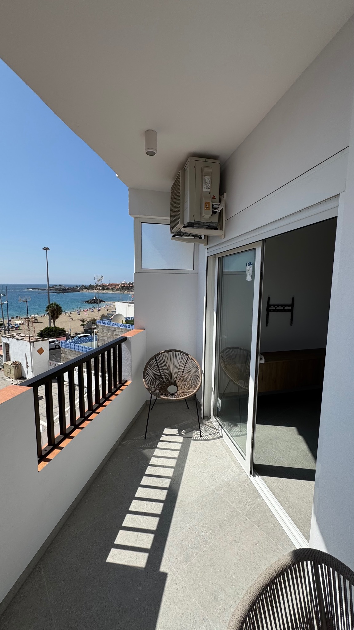 Jasny balkon z widokiem na plażę, szare płytki podłogowe, wiklinowe krzesła, klimatyzator na ścianie, czarne barierki.