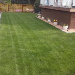 Zadbany trawnik z równo przystrzyżoną trawą, obrzeżem z kamienia i dekoracyjnymi donicami z kwiatami przy drewnianym tarasie. W tle ozdobne ogrodzenie.