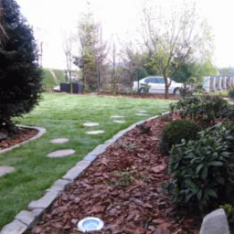 Zadbany ogród z równo przystrzyżonym trawnikiem, ścieżką z kamieni i rabatami wyłożonymi korą. W tle widoczny samochód i elementy architektury ogrodowej.