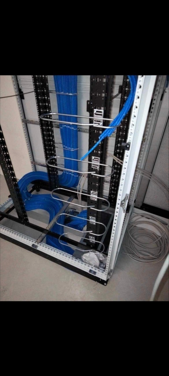 Ułożone niebieskie przewody w metalowej szafie rack, widoczne uchwyty kablowe i zwój szarych kabli na podłodze.
