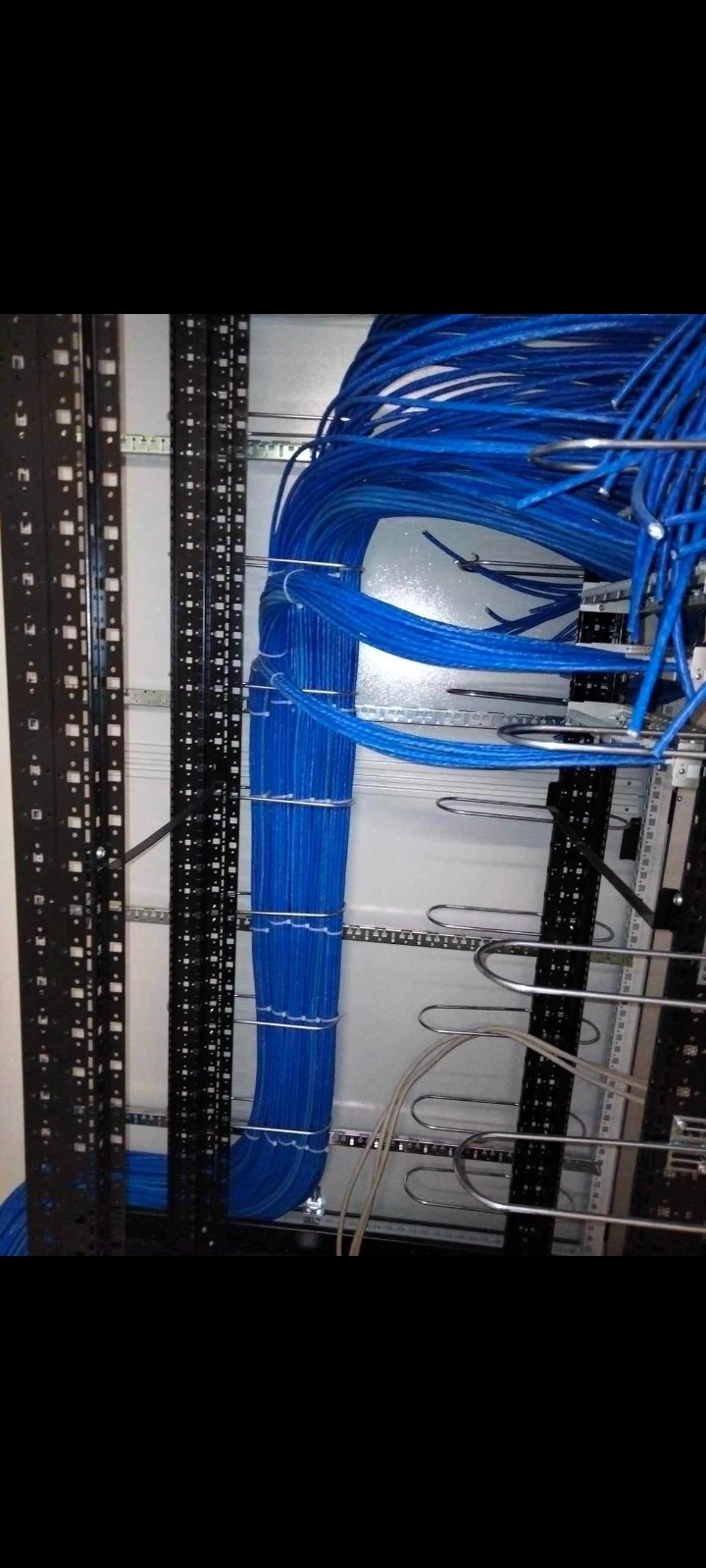 Pionowo ułożone, niebieskie kable telekomunikacyjne w szafie rack, uporządkowane za pomocą metalowych uchwytów, widoczne czarne szyny montażowe z otworami.