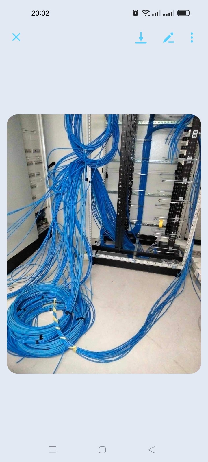 Niebieskie kable telekomunikacyjne zwinięte w krąg, częściowo rozwinięte, prowadzone do metalowej szafy rack z zamontowanymi panelami krosowniczymi.