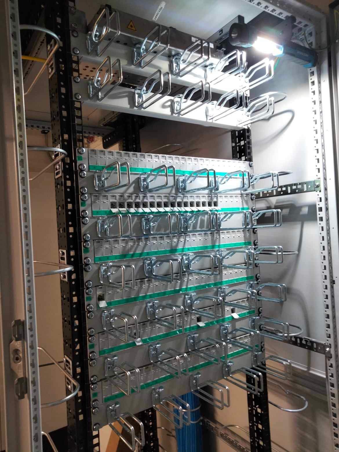 Widok z boku na szafę telekomunikacyjną z zamontowanymi uchwytami kablowymi i częściowo poprowadzonymi kablami w kolorze niebieskim, oświetloną od góry lampą inspekcyjną.