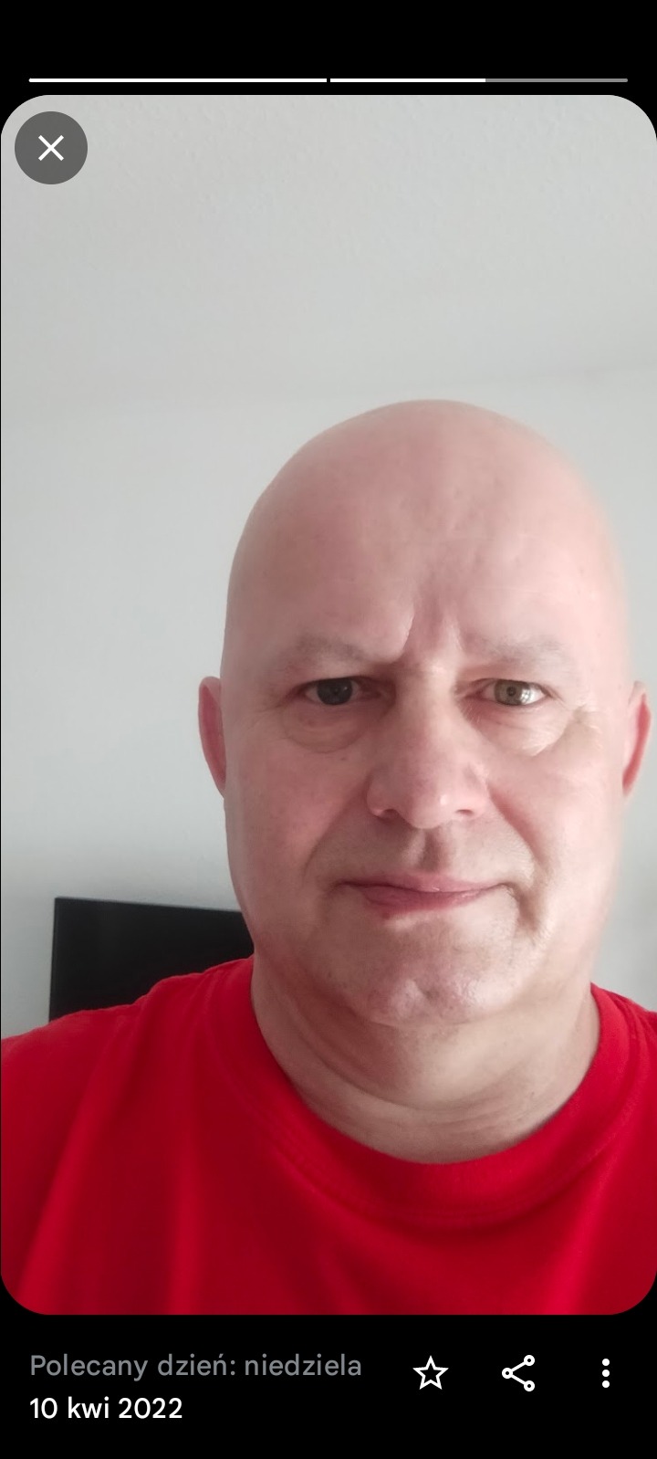 Portret mężczyzny w czerwonej koszulce z heterochromią, spoglądającego w obiektyw na tle jasnej ściany.