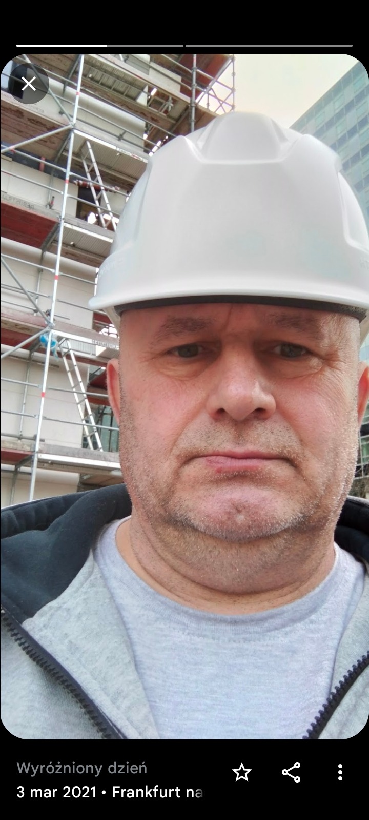 Mężczyzna w białym kasku ochronnym na tle rusztowania i budynku biurowego.