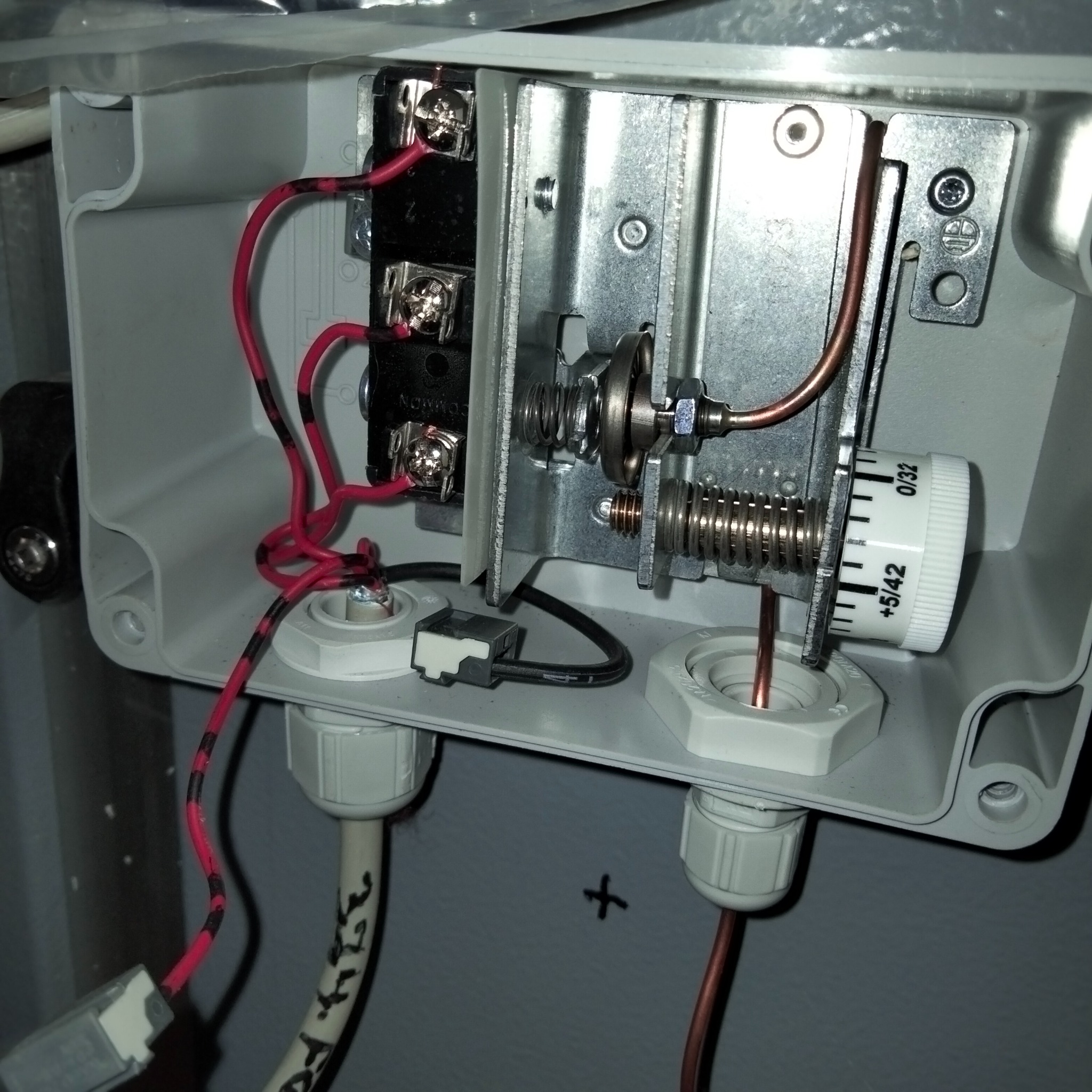 Wnętrze szarej skrzynki przyłączeniowej z metalowymi elementami, przewodami czerwono-czarnymi i miedzianymi, oraz białym pokrętłem regulacyjnym z podziałką.