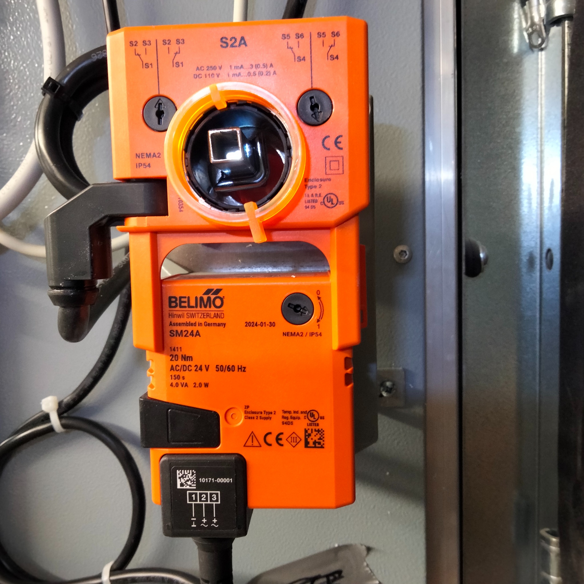Pomarańczowy siłownik Belimo SM24A z widocznymi parametrami technicznymi, zamontowany na metalowej szafie sterowniczej, z podłączonymi przewodami.