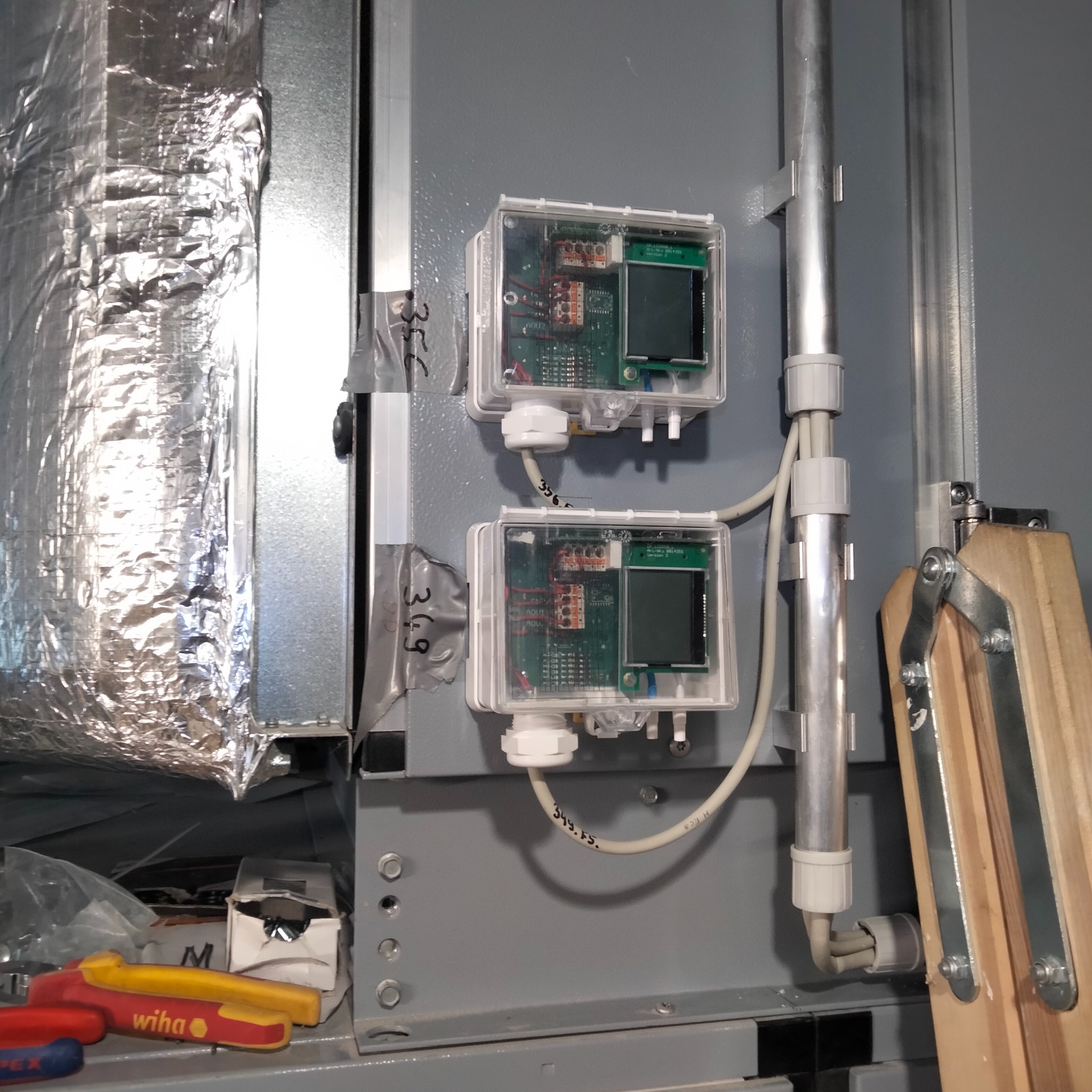 Dwa elektroniczne czujniki z wyświetlaczami zamontowane na szarej ścianie obok izolowanego przewodu wentylacyjnego, widoczne narzędzia Wiha i drabina.