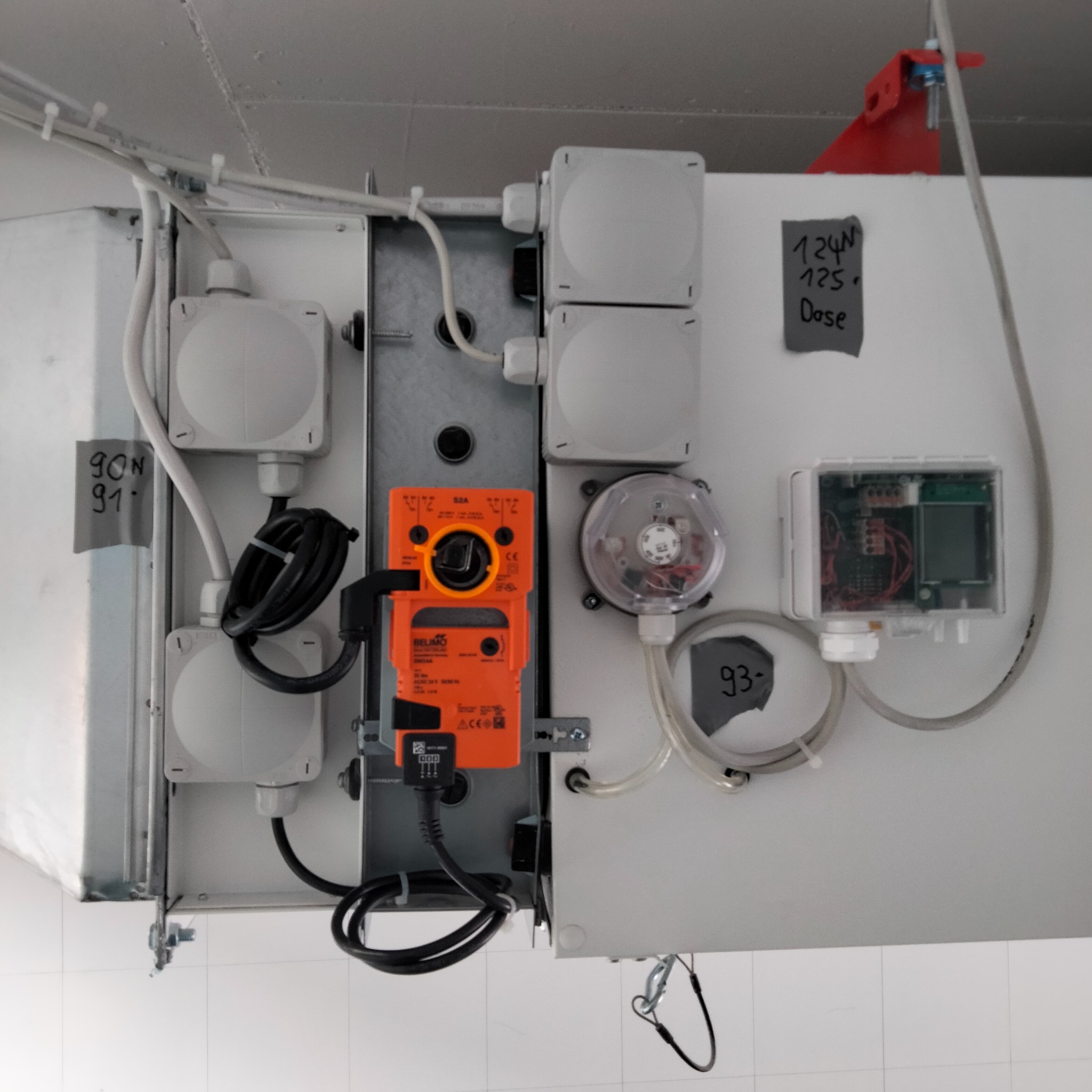 Szczegółowe ujęcie instalacji wentylacyjnej z czujnikami, przewodami i elementami sterującymi, w tym pomarańczowy siłownik Belimo, widoczne numery identyfikacyjne na taśmach.