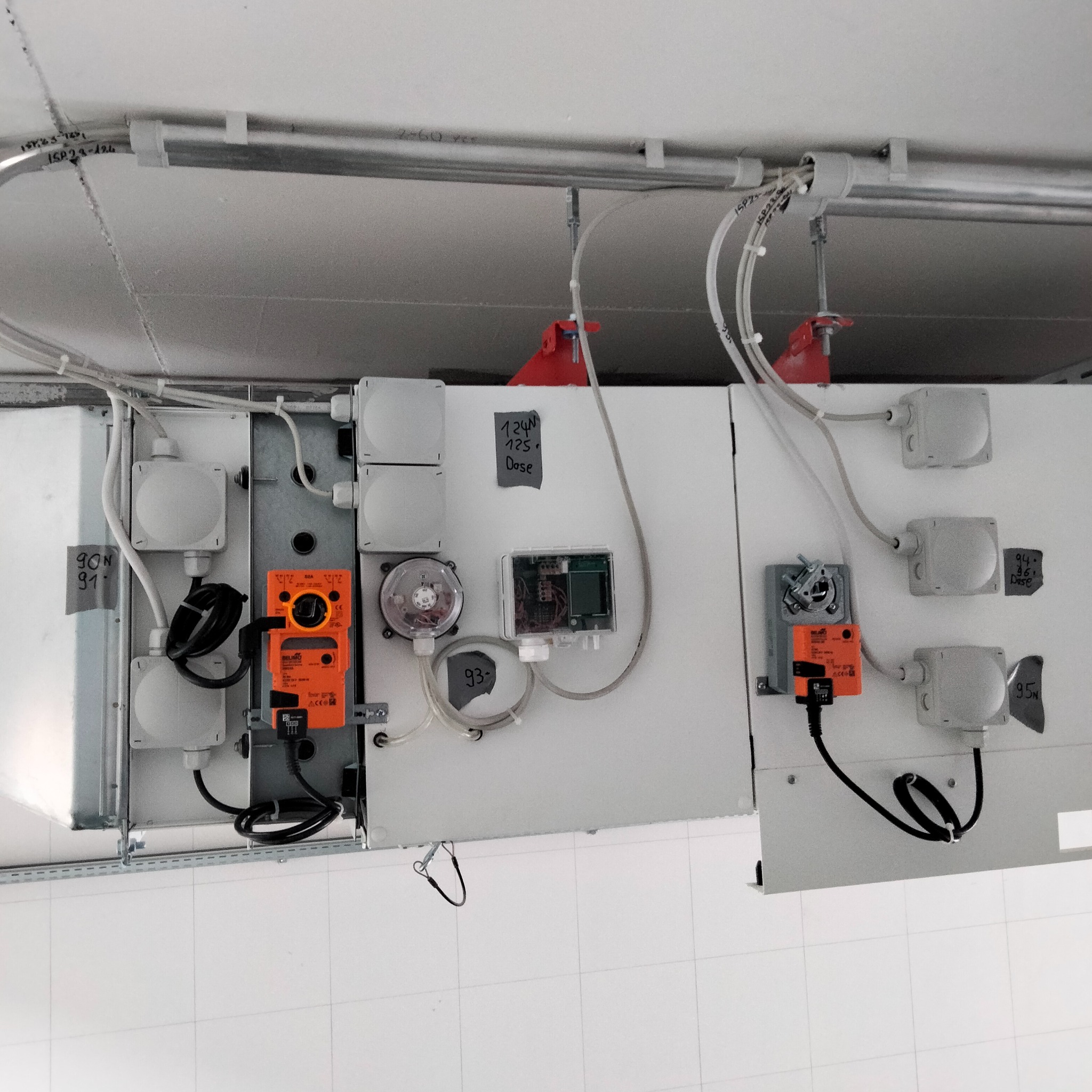 Szafa sterownicza z elementami automatyki, przewodami i czujnikami, zamontowana pod sufitem w pomieszczeniu z białą, kwadratową płytką na ścianach.