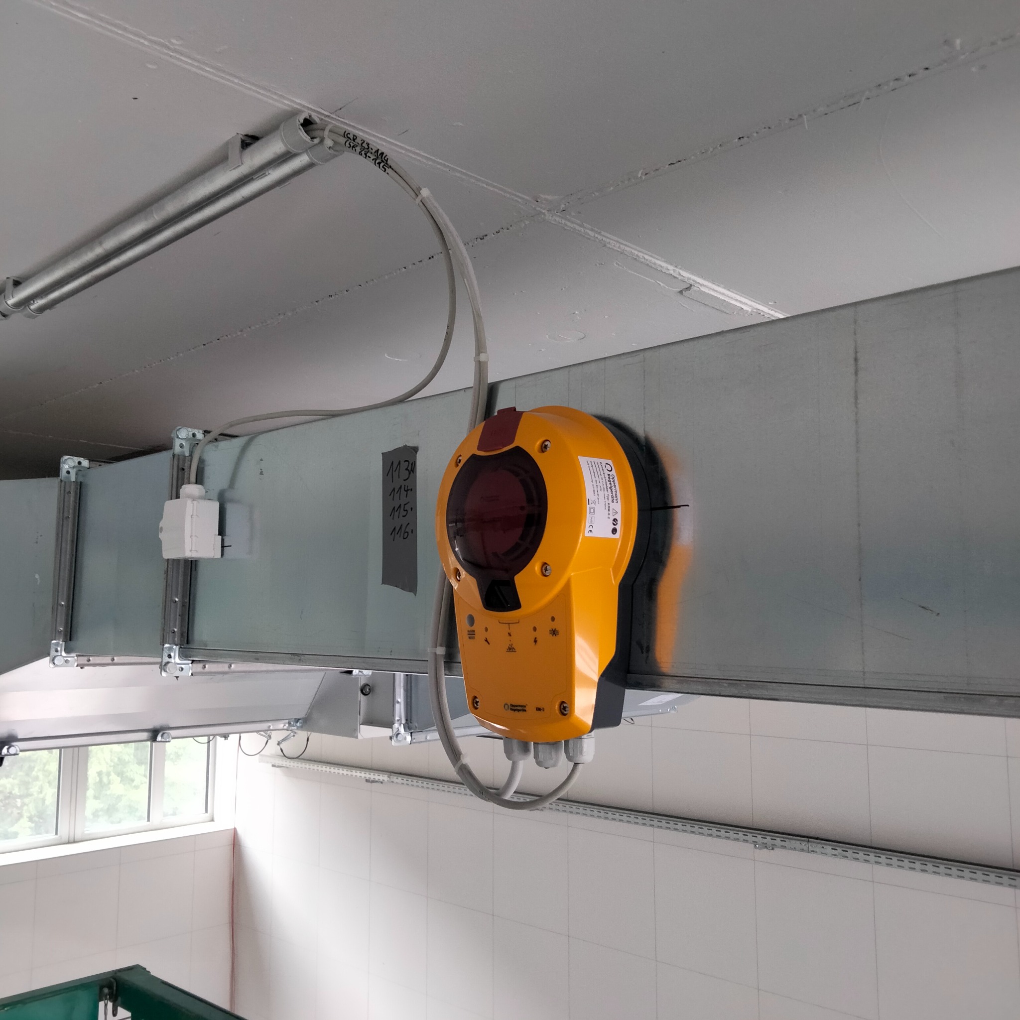 Żółty czujnik dymu zamontowany na metalowym kanale wentylacyjnym pod sufitem, widoczne przewody i oznaczenia na kanale.