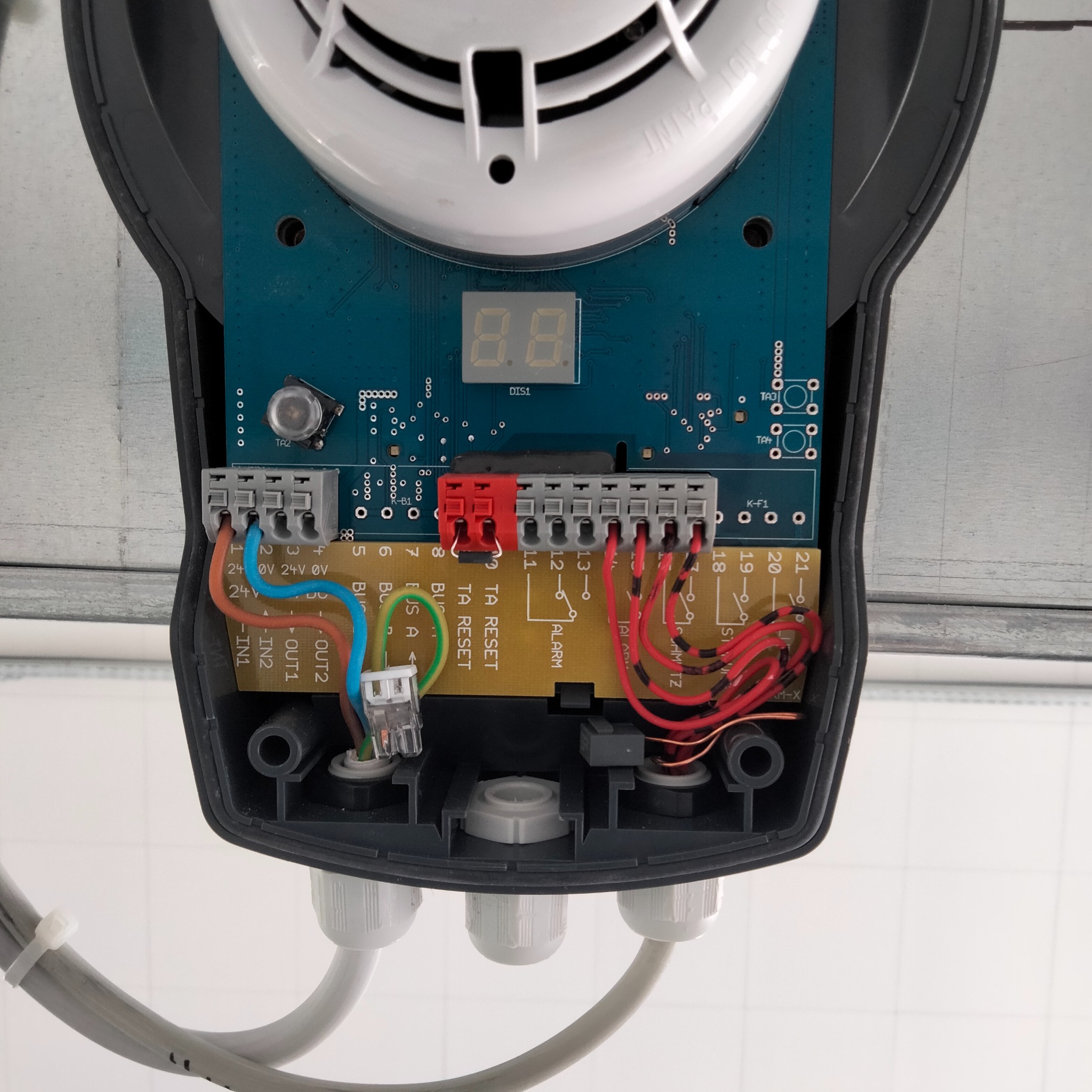 Otwarta obudowa sterownika bramy automatycznej z widoczną elektroniką, przewodami i złączkami. Widoczna płyta główna z wyświetlaczem, terminale podłączeniowe i kable w różnych kolorach.