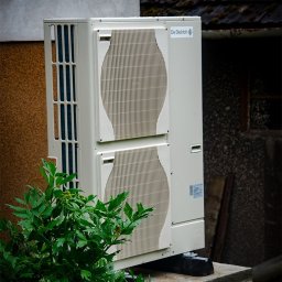 W.H GROUP SPÓŁKA Z OGRANICZONĄ ODPOWIEDZIALNOŚCIĄ - Zewnętrzna jednostka klimatyzacji De Dietrich zamontowana na betonowym fundamencie, częściowo zasłonięta przez bujną, zieloną roślinność. W tle widoczna ściana budynku z różnymi fakturami i dach...