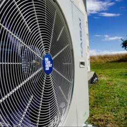 W.H GROUP SPÓŁKA Z OGRANICZONĄ ODPOWIEDZIALNOŚCIĄ - Zewnętrzna jednostka klimatyzacji Kaisai z widocznym wentylatorem i naklejką 'We care about air', na tle zielonej trawy i błękitnego nieba z drzewem, logo firmy w prawym górnym rogu.