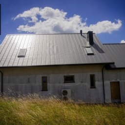 W.H GROUP SPÓŁKA Z OGRANICZONĄ ODPOWIEDZIALNOŚCIĄ - Nowoczesny dom jednorodzinny z szarą blachą na dachu, widocznym kominem, oknami dachowymi i klimatyzatorem na ścianie, otoczony wysoką trawą w słoneczny dzień.