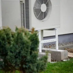 Zewnętrzna jednostka pompy ciepła De Dietrich zamontowana na białym stelażu, widoczna zza krzewu i rabaty z białymi kamykami.