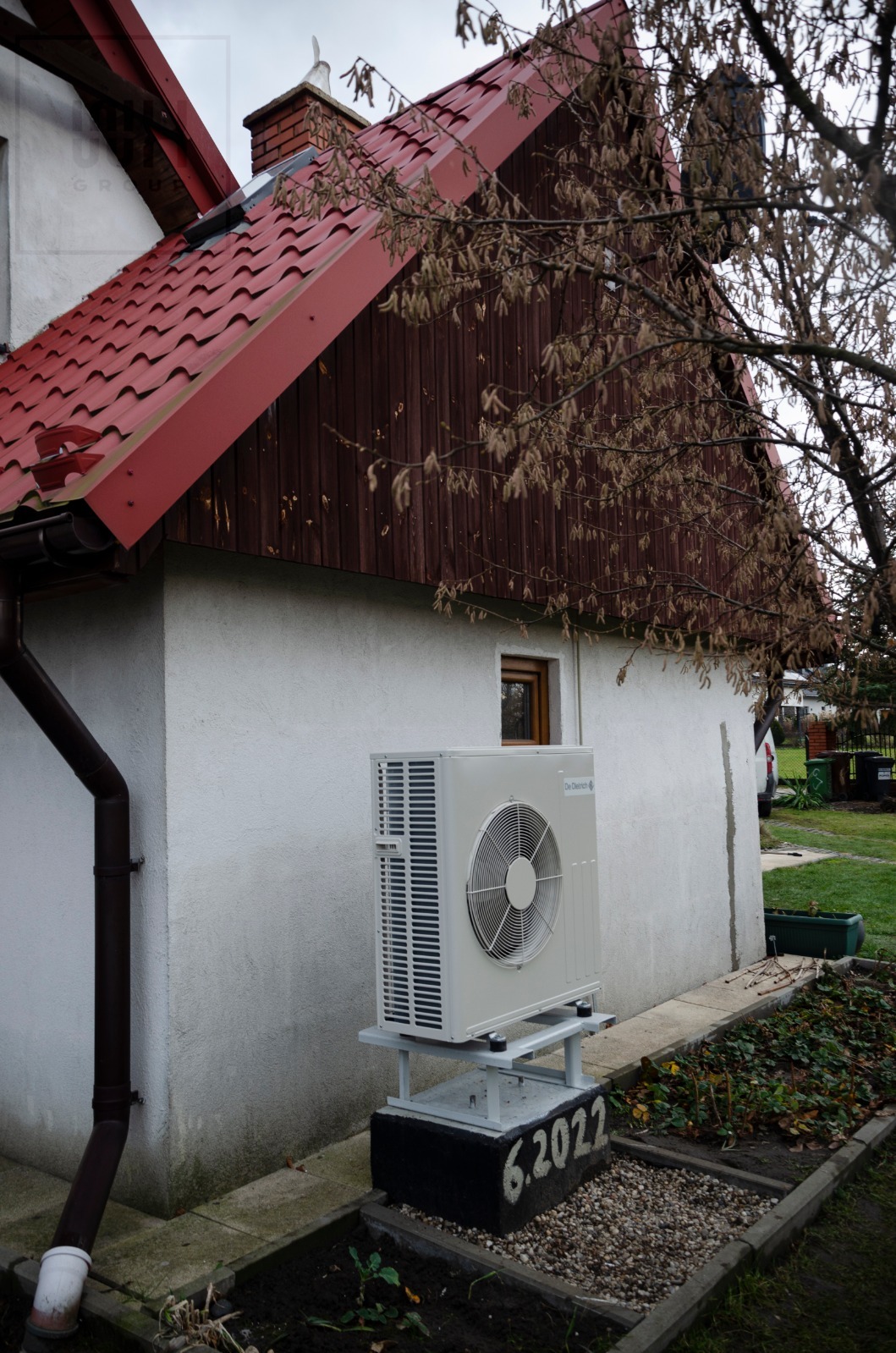 Biała pompa ciepła De Dietrich zamontowana na podwyższeniu z datą 6.2022, widoczna na tle białego domu z czerwoną dachówką i brązową boazerią, obok rynna i rabata.
