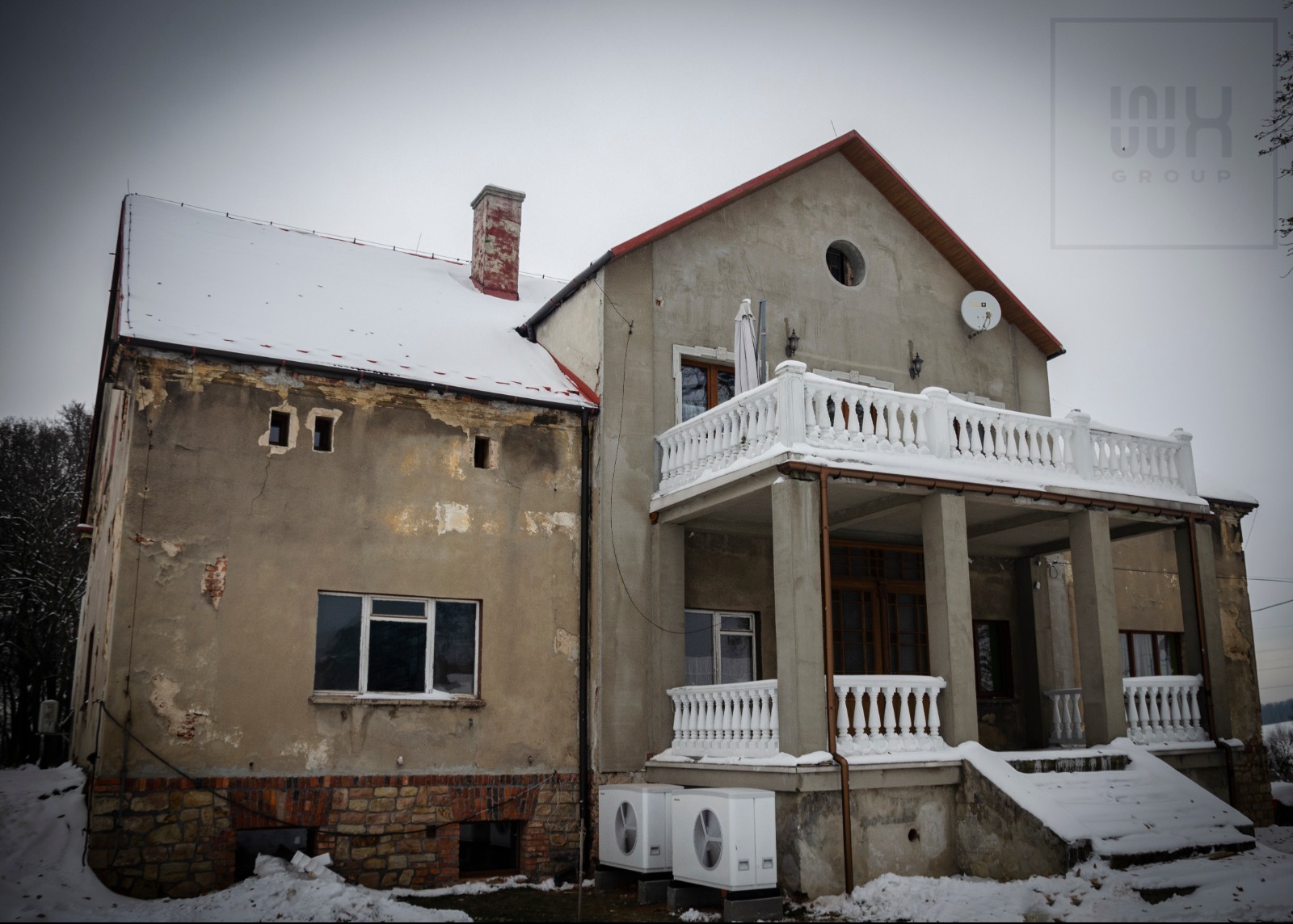 Dwukondygnacyjny dom z balkonem, pokryty śniegiem, z dwoma jednostkami zewnętrznymi klimatyzacji na zewnątrz. Starsza część budynku z widocznymi uszkodzeniami elewacji. Na dachu antena satelitarna.