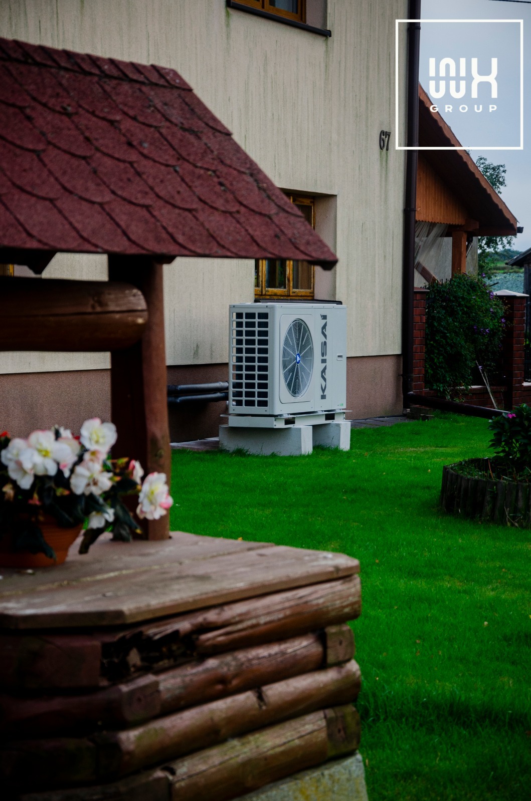 Zewnętrzna jednostka pompy ciepła Kaisai zamontowana obok domu z studnią, na zielonym trawniku, widoczny numer domu 67 oraz logo firmy instalacyjnej w rogu.