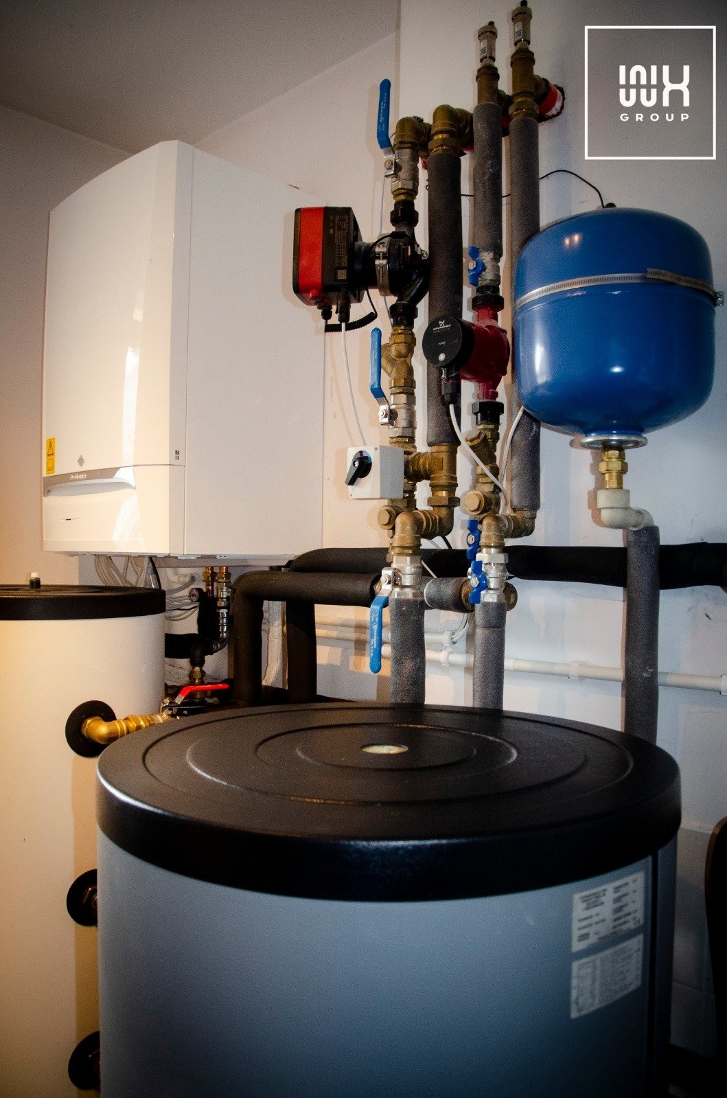 Instalacja z białym zasobnikiem ciepłej wody, szarym akumulatorem ciepła, niebieskim naczyniem przeponowym i licznymi rurami z zaworami w kotłowni