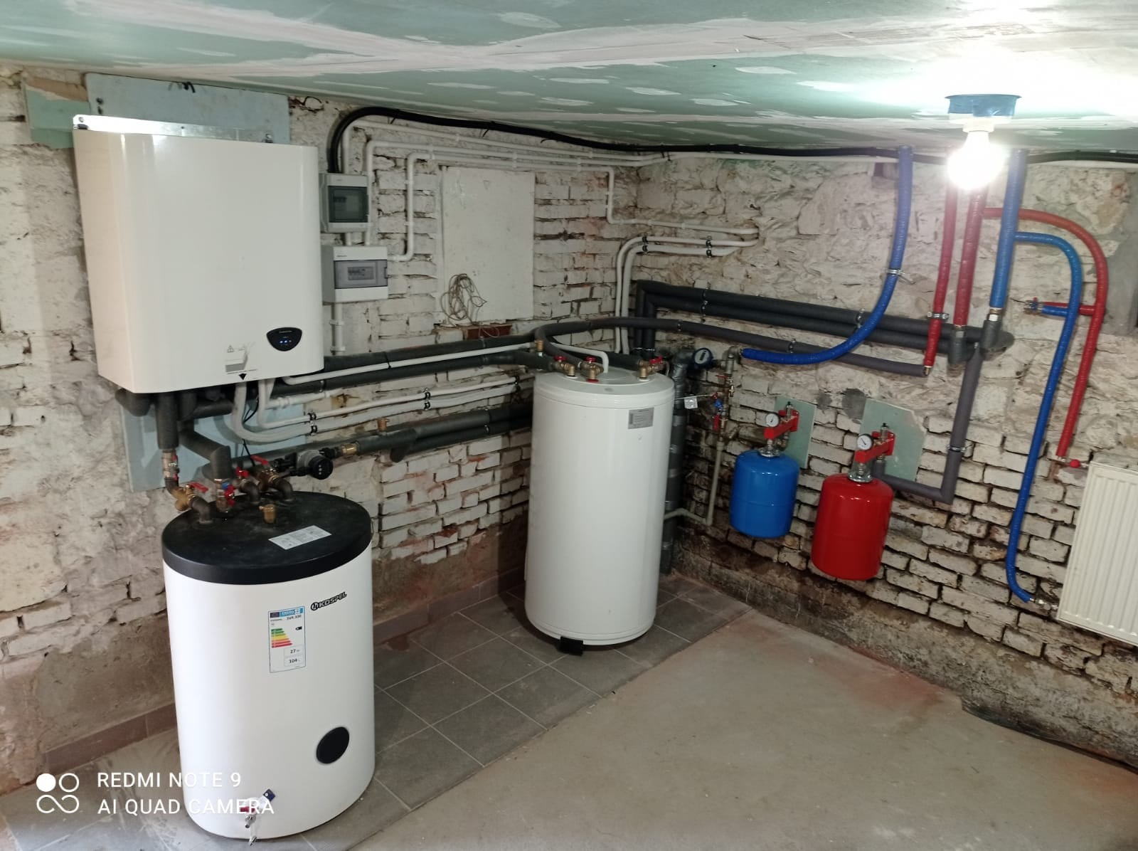 Dwa białe zasobniki ciepłej wody użytkowej, wiszący bojler, rury w izolacji, instalacja z czerwonymi i niebieskimi elementami w piwnicy z ceglanymi ścianami i surowym wykończeniem sufitu.