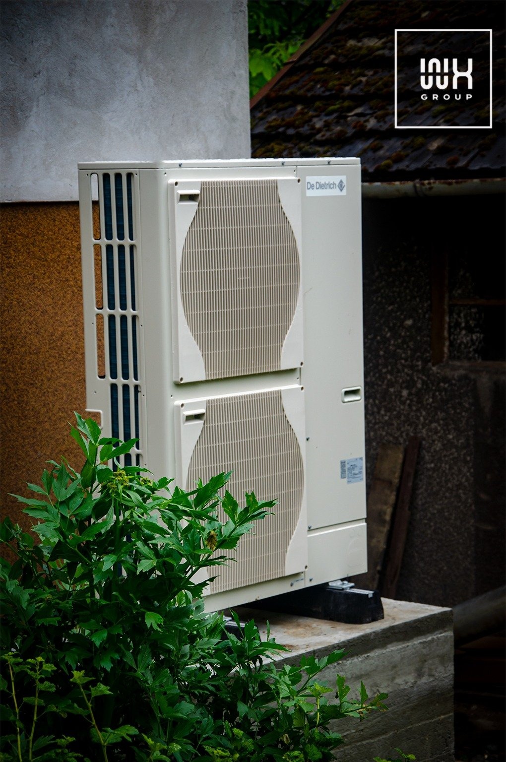 Zewnętrzna jednostka klimatyzacji De Dietrich zamontowana na betonowym fundamencie, częściowo zasłonięta przez bujną, zieloną roślinność. W tle widoczna ściana budynku z różnymi fakturami i dach...