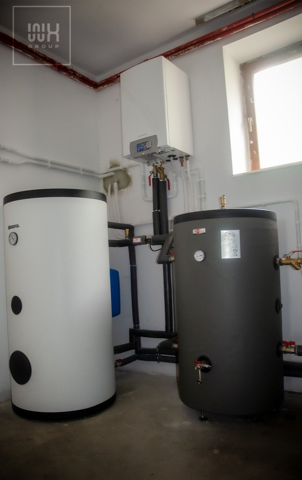 Biały i szary zbiornik buforowy podłączone do kotła grzewczego w pomieszczeniu z oknem, rury wzdłuż ścian.